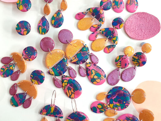 Photo of a collection of colourful and funky polymer clay earrings. From studs to big dangly earrings. They are multicolor but mainly have pops of pink and golden yellow. Material polymer clay with resin top coat.