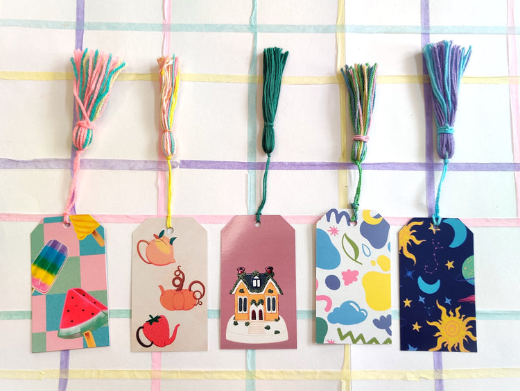 A set of five bookmarks are placed on a bright background. They are rectangular with handmade multicolor tassels. The themes of the bookmarks are pastel icecream, fruit teapots, cozy cottage, pastel doodles and astronomical designs.